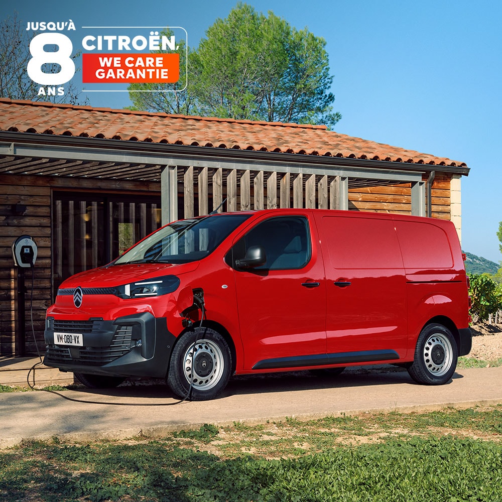Side three‑quarter exterior view of a red Citroën ë‑Jumpy charging in front of a building, showing modern electric design, everyday practicality, connectivity and confident professional presence.