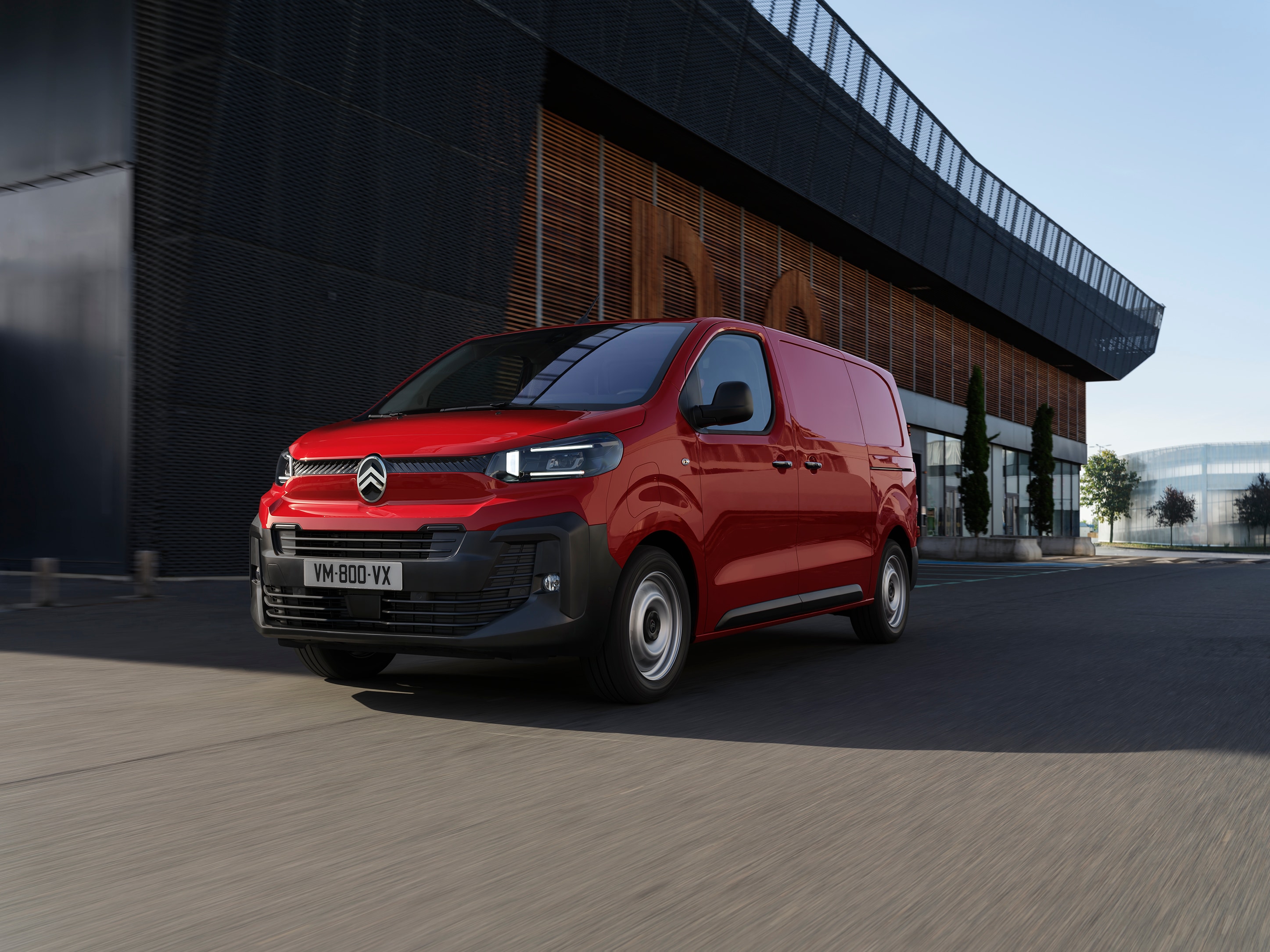Vue extérieure de trois quarts avant d'une Citroën Jumpy rouge garée près d'un bâtiment moderne, mettant en valeur son design professionnel, son aspect pratique et sa présence assurée.