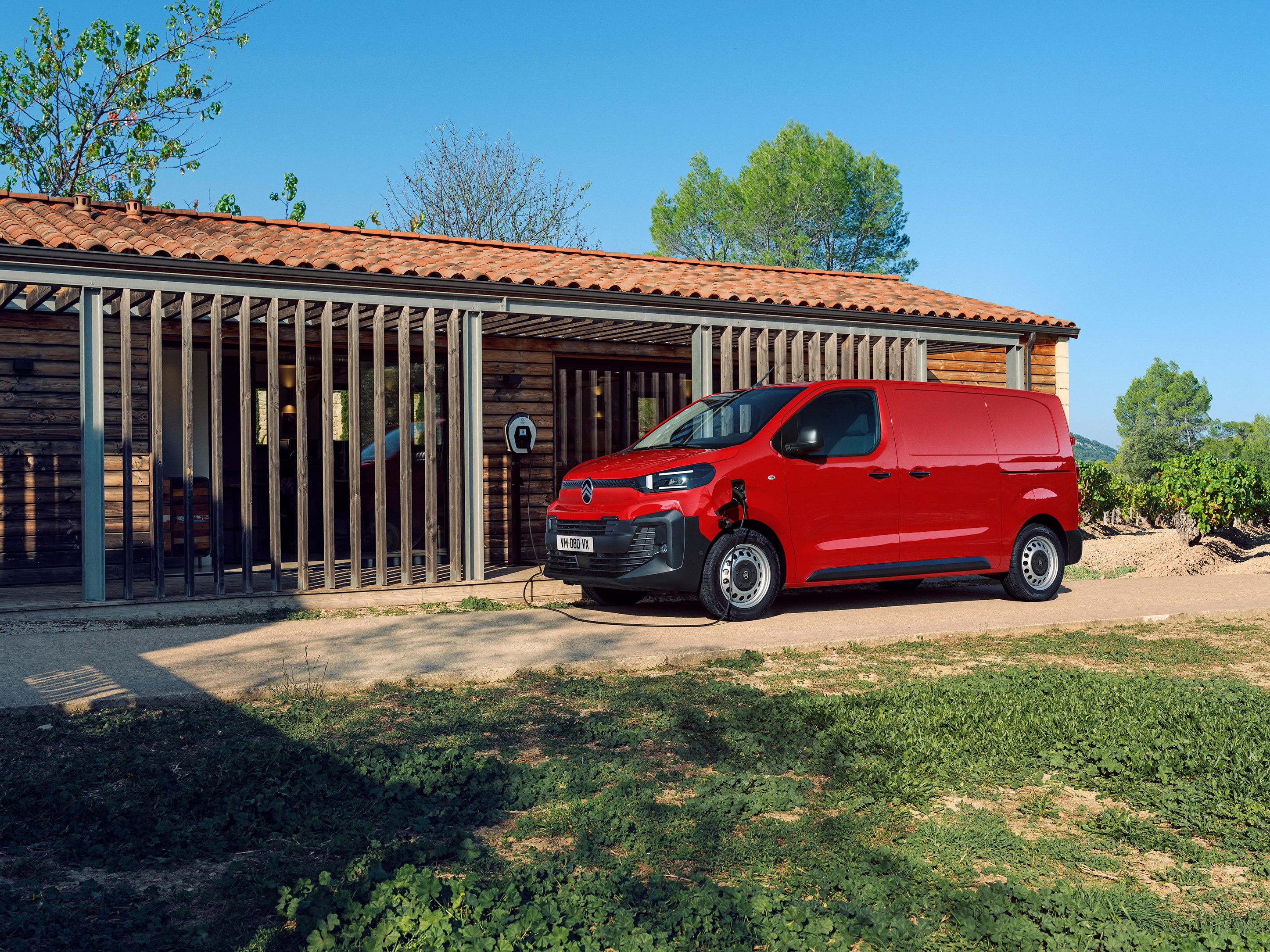 Vue extérieure de trois quarts d'un Citroën ë-Jumpy rouge en charge devant un bâtiment, mettant en avant la mobilité électrique, la praticité au quotidien et une présence professionnelle assurée.