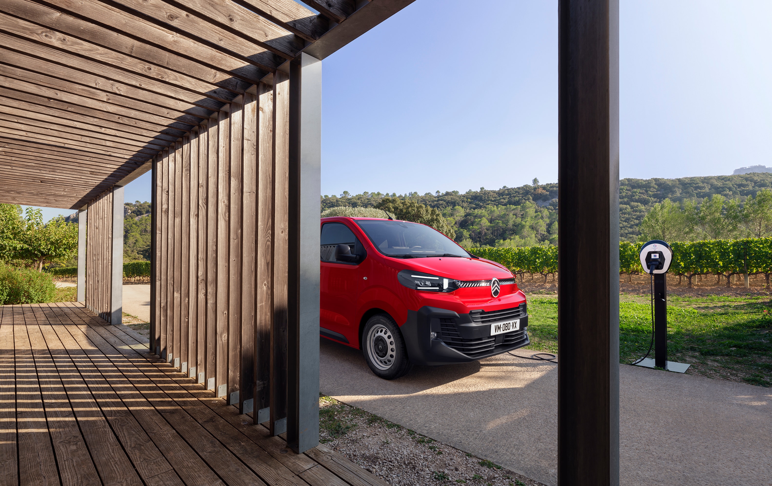 Vue extérieure de profil trois-quarts d'une Citroën ë-Jumpy rouge garée devant un bâtiment en bois, mettant en avant le design électrique moderne, la praticité au quotidien et une présence professionnelle assurée.