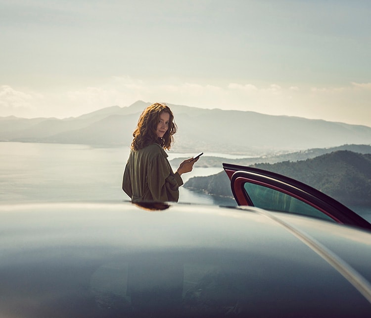 زن در پس زمینه وسیله نقلیه ای که به تلفن هوشمند خود در فضای باز نگاه می کند